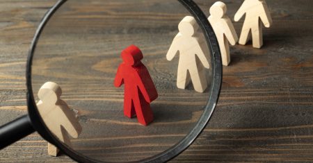 Magnifying glass focuses on a red wooden figure from the crowd of wooden figures on a wooden table. Leadership concept