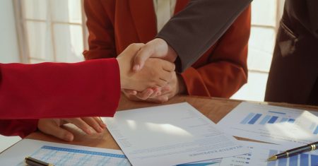 Indoor business meeting in a modern office. Confident businesswoman and businessman shaking hands. Young professionals working with laptops, discussing strategies, and making connections.
